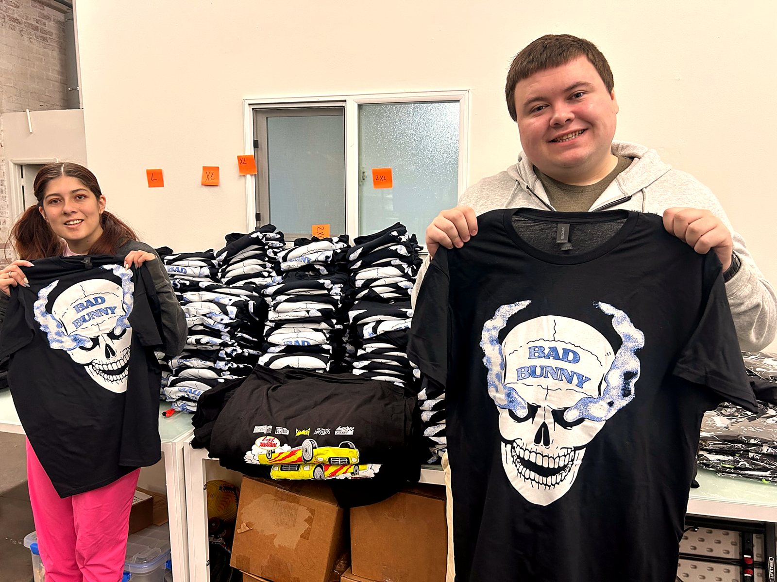 Two participants holding up finished screen-printed shirts beside a stack of completed order